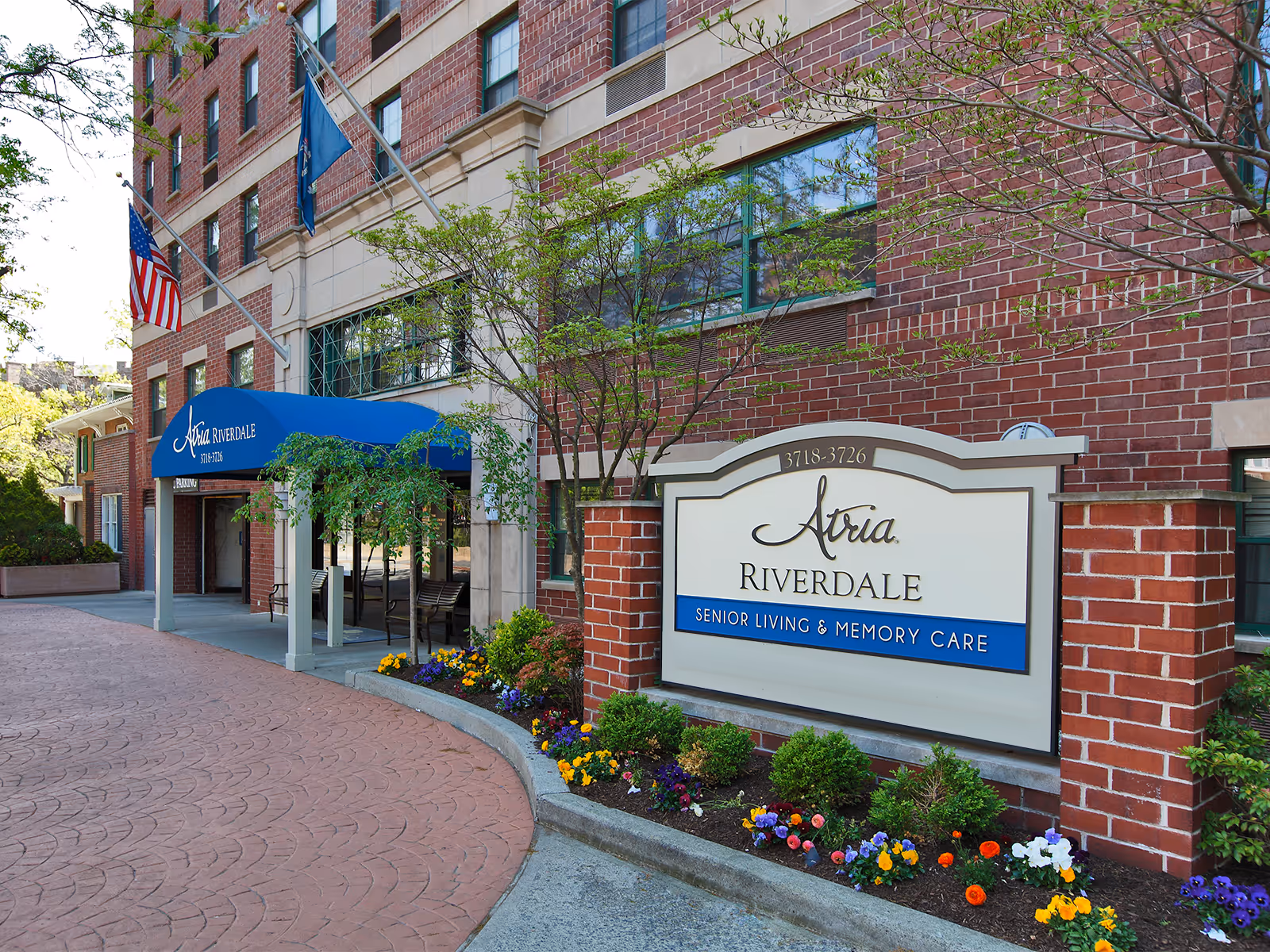 Exterior view of Atria Riverdale senior living and memory care facility with a brick facade, blue awning over the entrance, American and state flags, and a landscaped flower bed in front of the building sign.