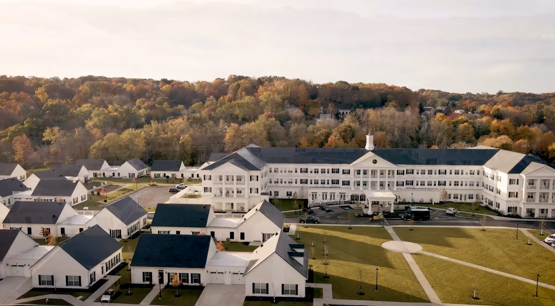 Aerial view of Ashton Commons senior living facility showing multiple white residential buildings with dark roofs, a large three-story main building, parking areas, and green lawns with pathways, set against a backdrop of trees with autumn foliage.