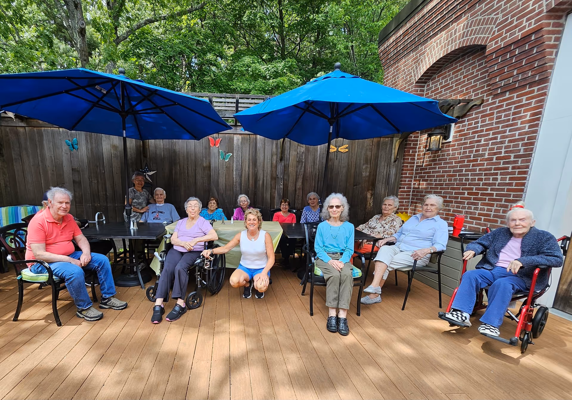 A group of elderly people sitting outdoors on a wooden deck under two large blue umbrellas. They are seated on chairs and wheelchairs, arranged in a semi-circle, with a woman kneeling in front of them. The background features a wooden fence decorated with colorful butterfly ornaments and a brick wall on the right side. Trees and greenery are visible behind the fence.