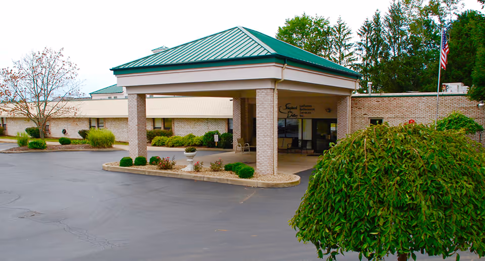 Entrance of Shepherd of the Valley - Boardman facility showing a covered drop-off area with a green metal roof, brick pillars, and surrounding landscaping including bushes and trees. An American flag is visible on the right side near the building.
