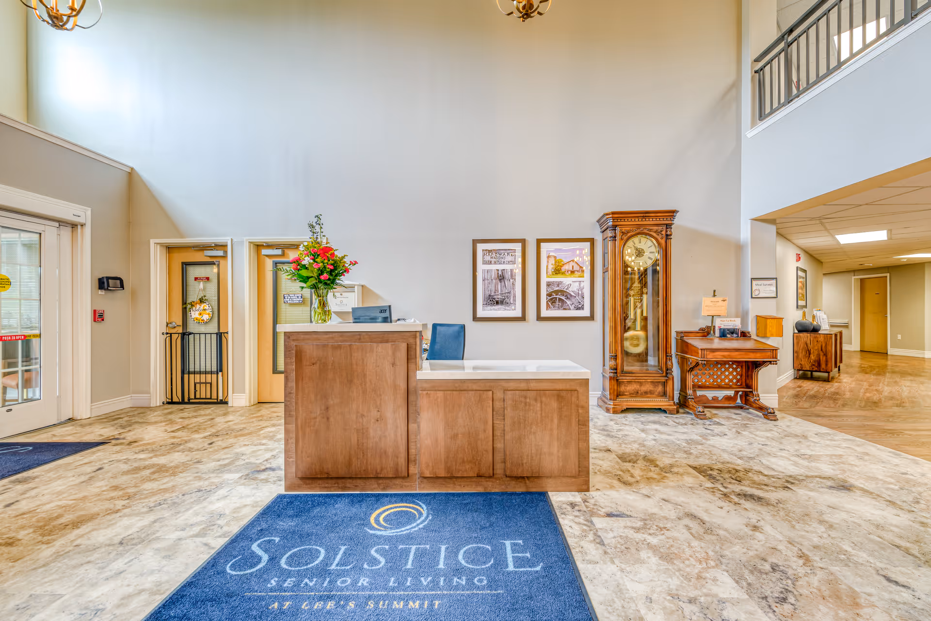 Reception area of Solstice Senior Living at Lee's Summit featuring a wooden front desk with a vase of flowers, a blue chair, a grandfather clock, framed pictures on the wall, and a blue floor mat with the facility's name. The space has high ceilings, beige walls, and a tiled floor.