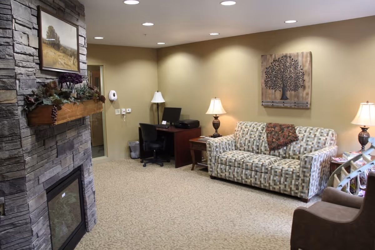 A cozy living room area with a patterned sofa, a brown armchair, two table lamps on side tables, a wooden desk with a computer and office chair, and a stone fireplace decorated with a painting and greenery.