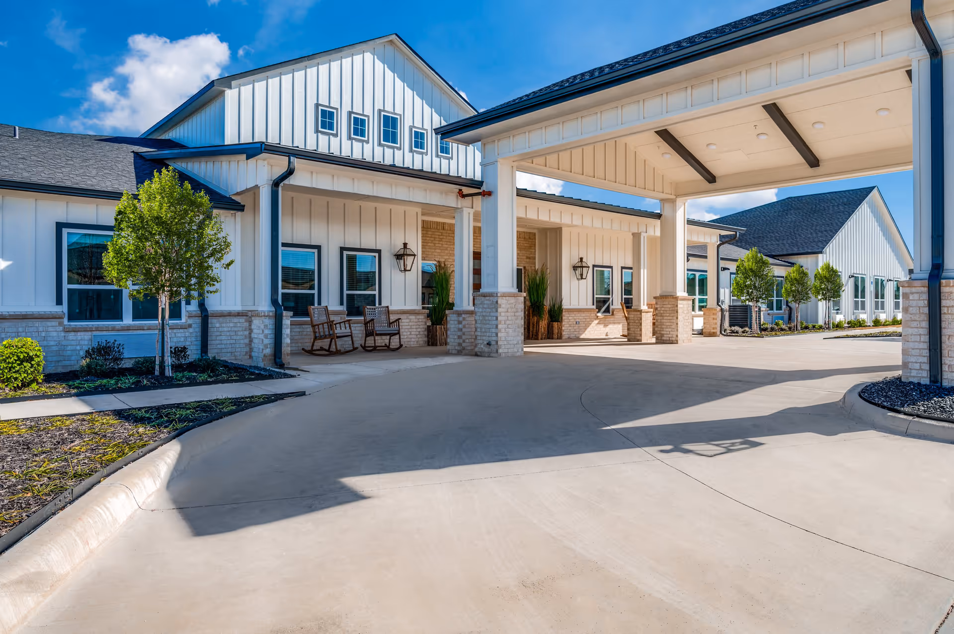 Exterior view of The Lodge of Saginaw Health & Wellness showing a covered entrance with brick and white panel siding, several windows, outdoor rocking chairs, small trees, and a clear blue sky.