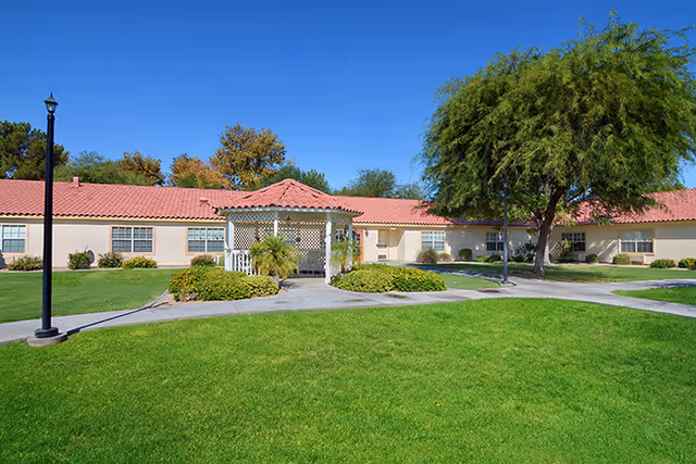Single-story building with a red tile roof and beige walls, surrounded by green grass, shrubs, and trees under a clear blue sky. A white gazebo is centered in front of the building, with paved walkways leading to it and a black lamppost on the left side.