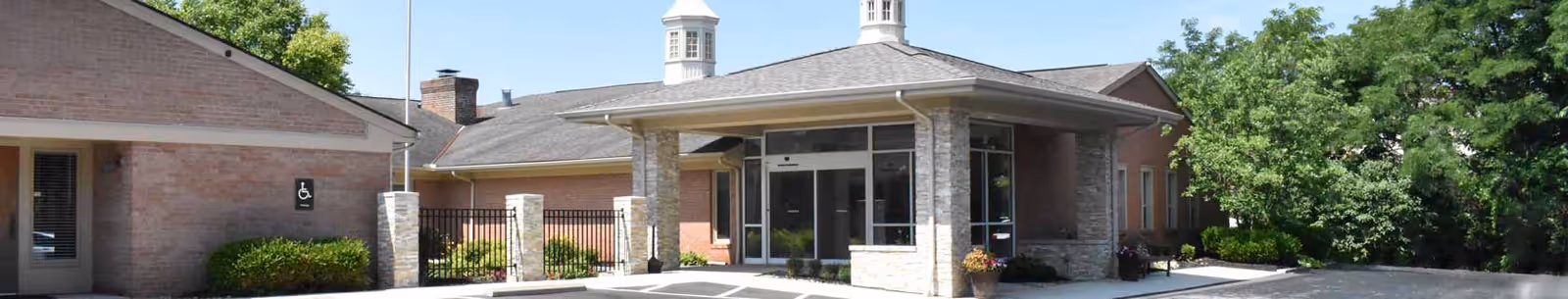 Front exterior view of Willow Brook Christian Home showing a single-story brick building with a covered entrance supported by stone pillars, surrounded by greenery and trees under a clear sky.