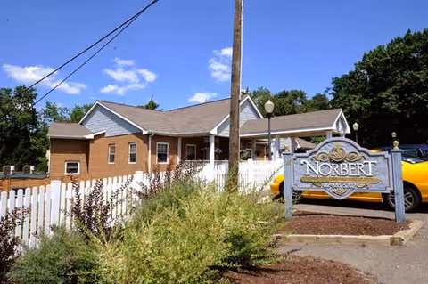 Exterior view of Norbert Personal Care facility showing a brick and siding building with a white picket fence, a garden area with shrubs, a yellow car parked nearby, and a sign with the facility name and phone number under a clear blue sky.
