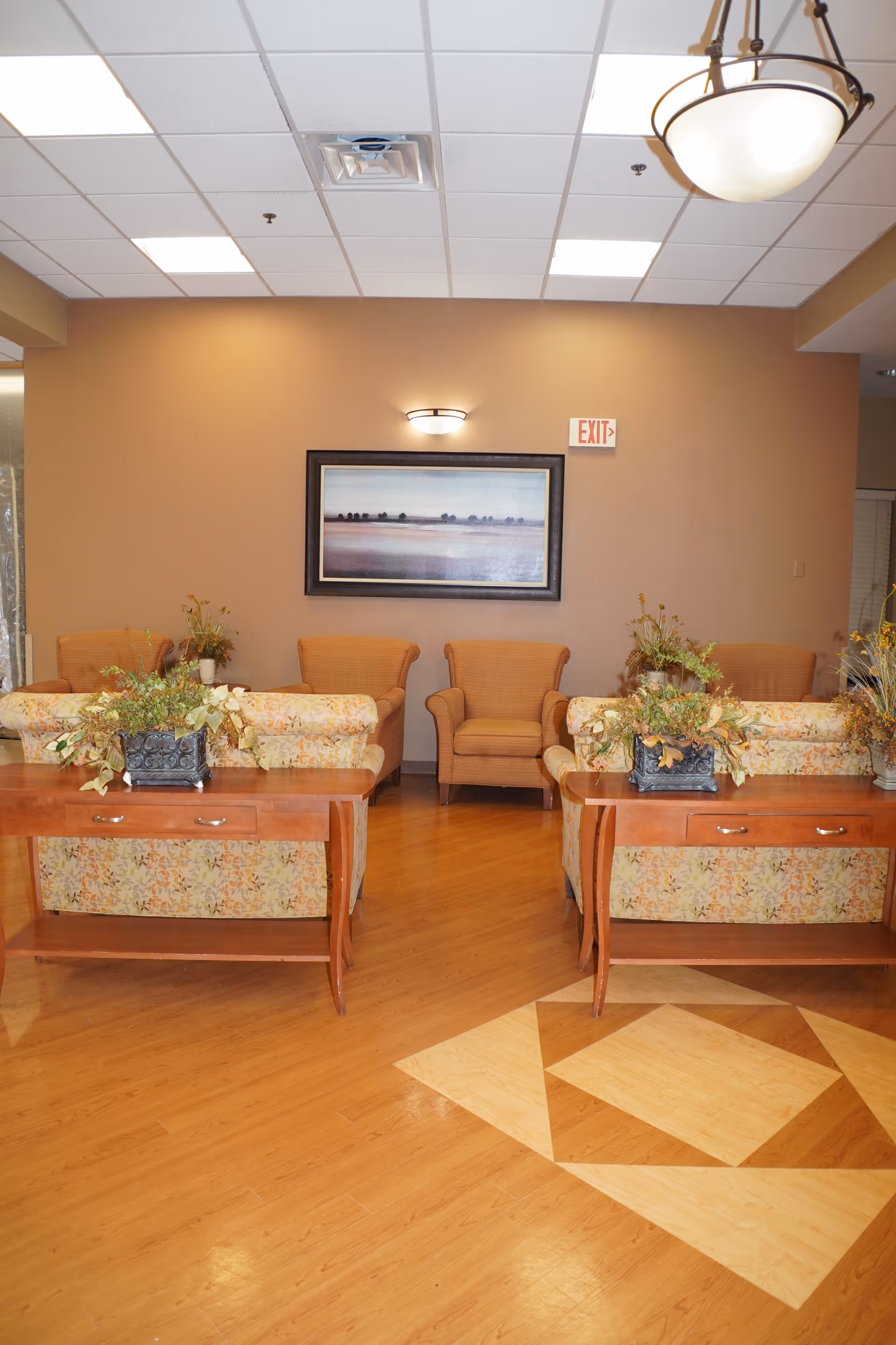 A cozy seating area in a senior living facility with floral patterned sofas, wooden side tables with decorative plants, and comfortable armchairs against a beige wall. A framed landscape painting and an exit sign are visible on the wall, with a ceiling light fixture above.