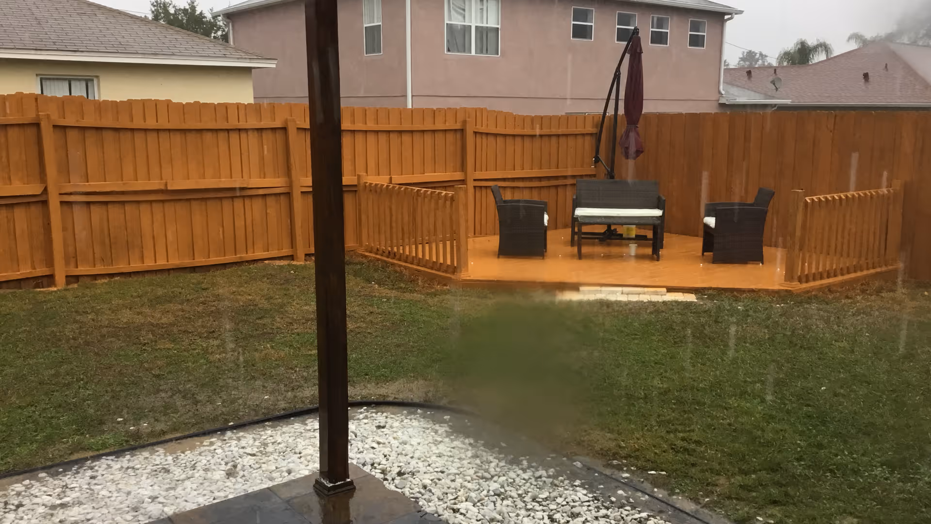 Outdoor fenced backyard area with a wooden deck featuring outdoor seating including two chairs, a loveseat, and a closed umbrella. The yard has grass and a small section with white decorative stones near a wooden post.