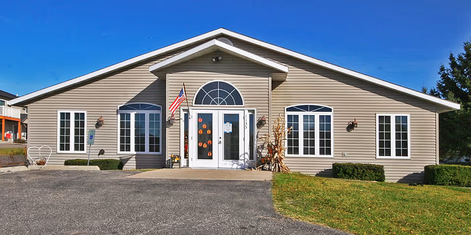 Exterior front view of a single-story beige building with white trim, featuring a central entrance with double glass doors decorated with Halloween pumpkin stickers, an American flag mounted beside the door, and some autumn decorations including cornstalks. The building is surrounded by a paved driveway and green grass lawn under a clear blue sky.