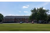 A large, two-story brick building with multiple windows, situated behind a wide grassy lawn under a partly cloudy sky. Several cars are parked in front of the building, and there are a few trees near the parking area.