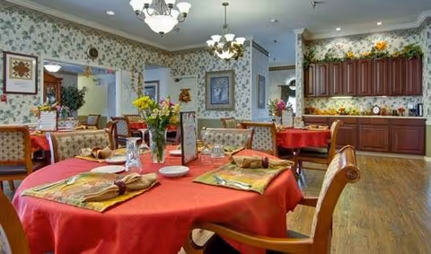 A dining room with tables covered in red tablecloths, each set with napkins, glasses, and menus. The room features floral wallpaper, wooden chairs with cushioned seats, and a wooden floor. There are chandeliers hanging from the ceiling and a sideboard with cabinets and decorative plants along the back wall.