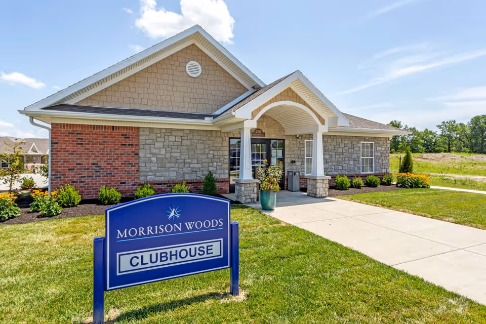 Exterior view of a single-story clubhouse building at Morrison Woods Health Campus. The building features a combination of brick and stone siding with a gabled roof and a covered entrance supported by columns. In front of the building, there is a blue sign that reads 'Morrison Woods Clubhouse' on a well-maintained lawn with landscaping and a concrete walkway leading to the entrance. The sky is clear with a few clouds.