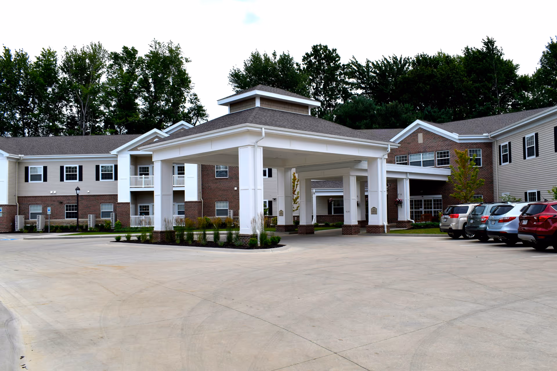 Exterior view of Danbury Mentor senior living facility showing a large covered entrance with white columns, surrounded by a two-story building with brick and beige siding. Several parked cars are visible on the right side, and trees are in the background.