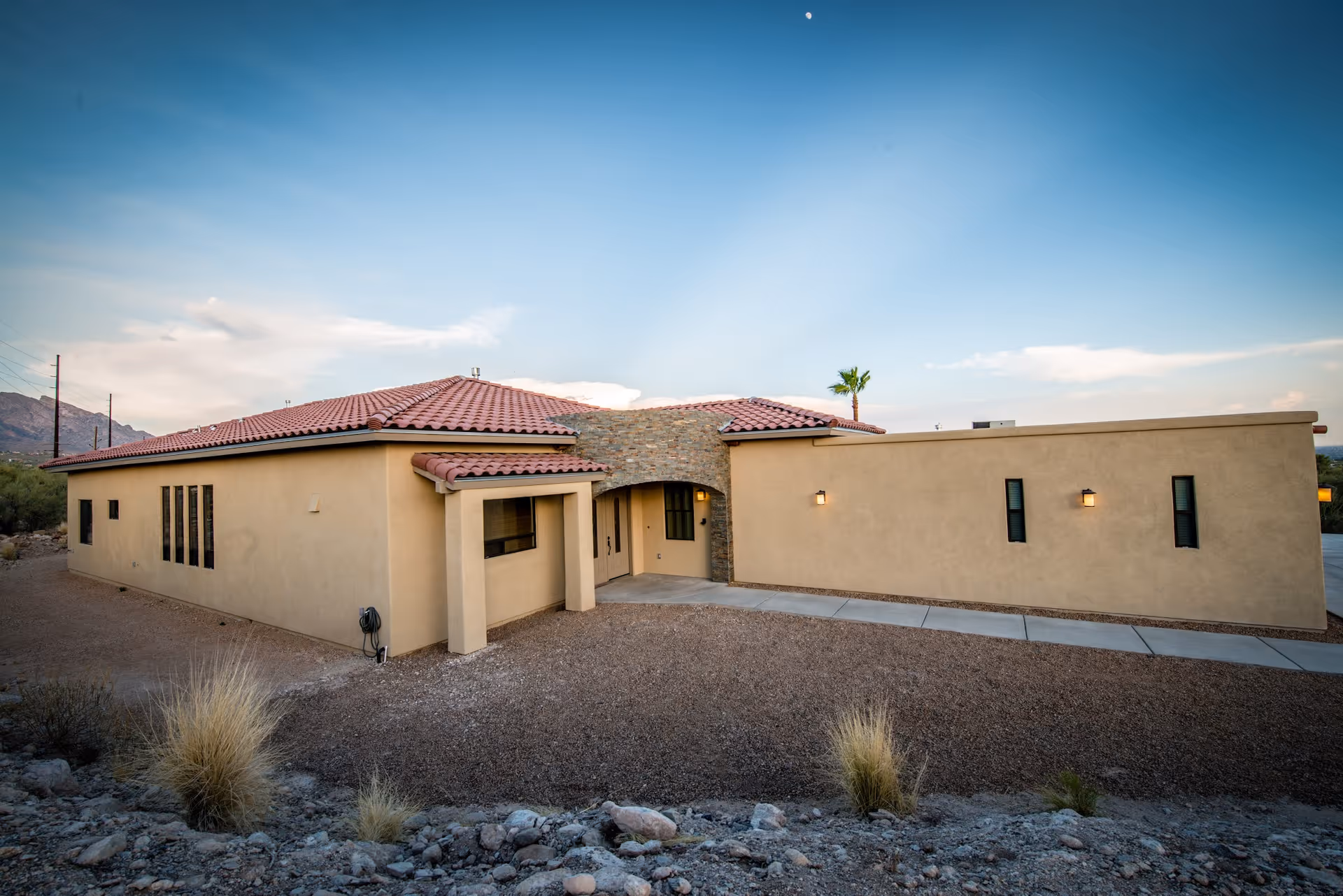 Exterior view of a single-story building with beige stucco walls and a red tile roof, set against a clear sky with a few clouds and a visible moon. The building has several windows and a stone archway entrance, surrounded by a gravel landscape with sparse desert vegetation.