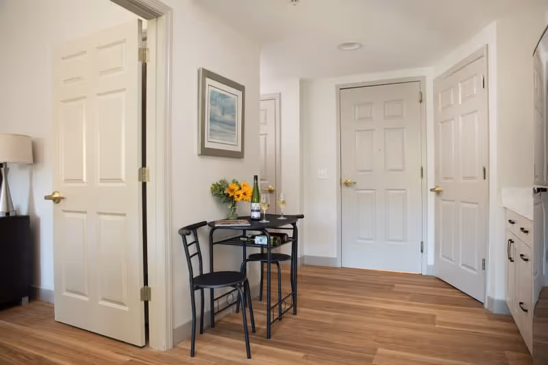 A small dining area in a senior living facility with a black metal table and two matching chairs. On the table are a bottle of wine, two wine glasses, a magazine, and a vase with yellow flowers. The room has light-colored walls, wooden flooring, and several white doors with gold handles. A framed picture hangs on the wall above the table.