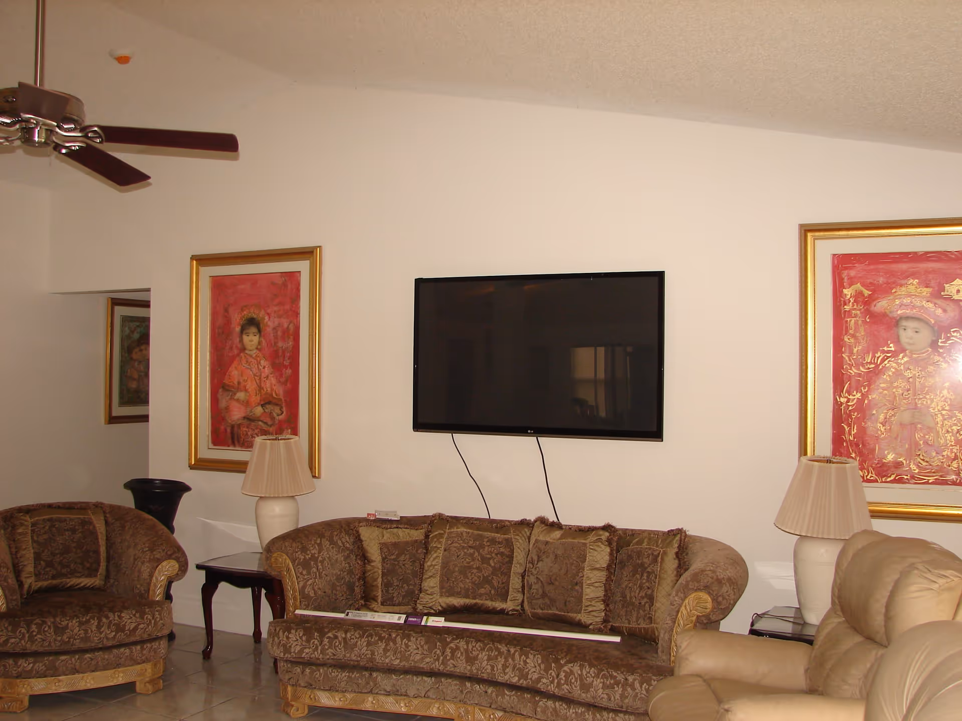 A living room with a brown patterned sofa and matching armchair, a beige recliner, two table lamps with beige shades on side tables, a ceiling fan with dark wooden blades, and a flat-screen TV mounted on a white wall. Two framed paintings with red backgrounds hang on the wall on either side of the TV.