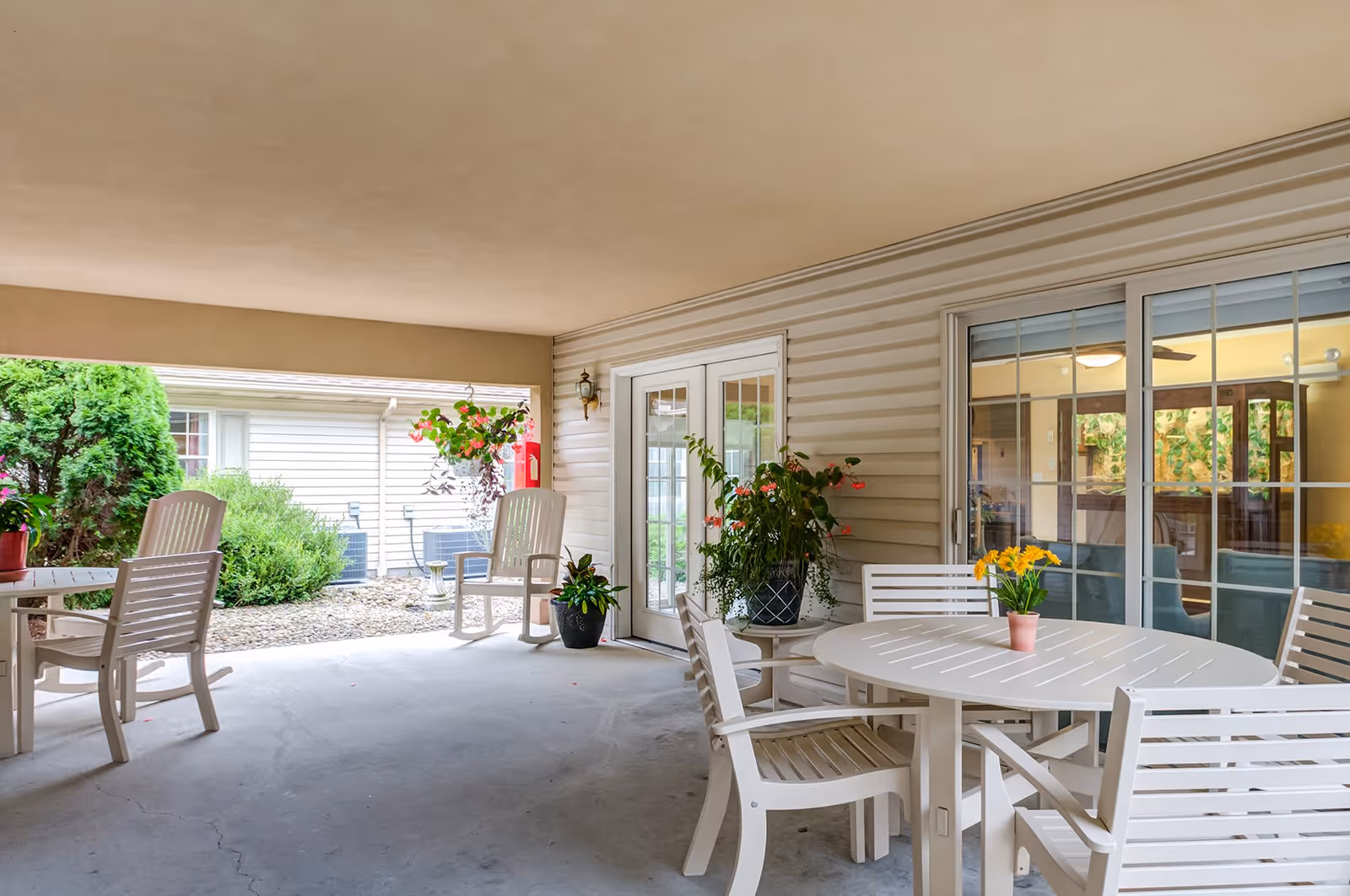 Covered outdoor patio area with white round table and chairs, potted plants, hanging flower basket, and sliding glass doors leading inside. Green bushes and another building are visible outside.