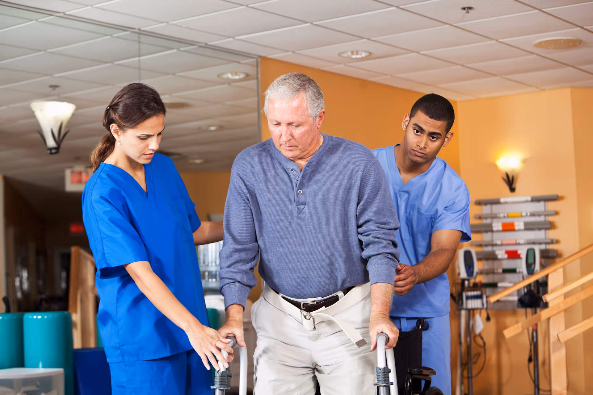 An elderly man using a walker is being assisted by two healthcare workers in blue scrubs inside a rehabilitation facility hallway.