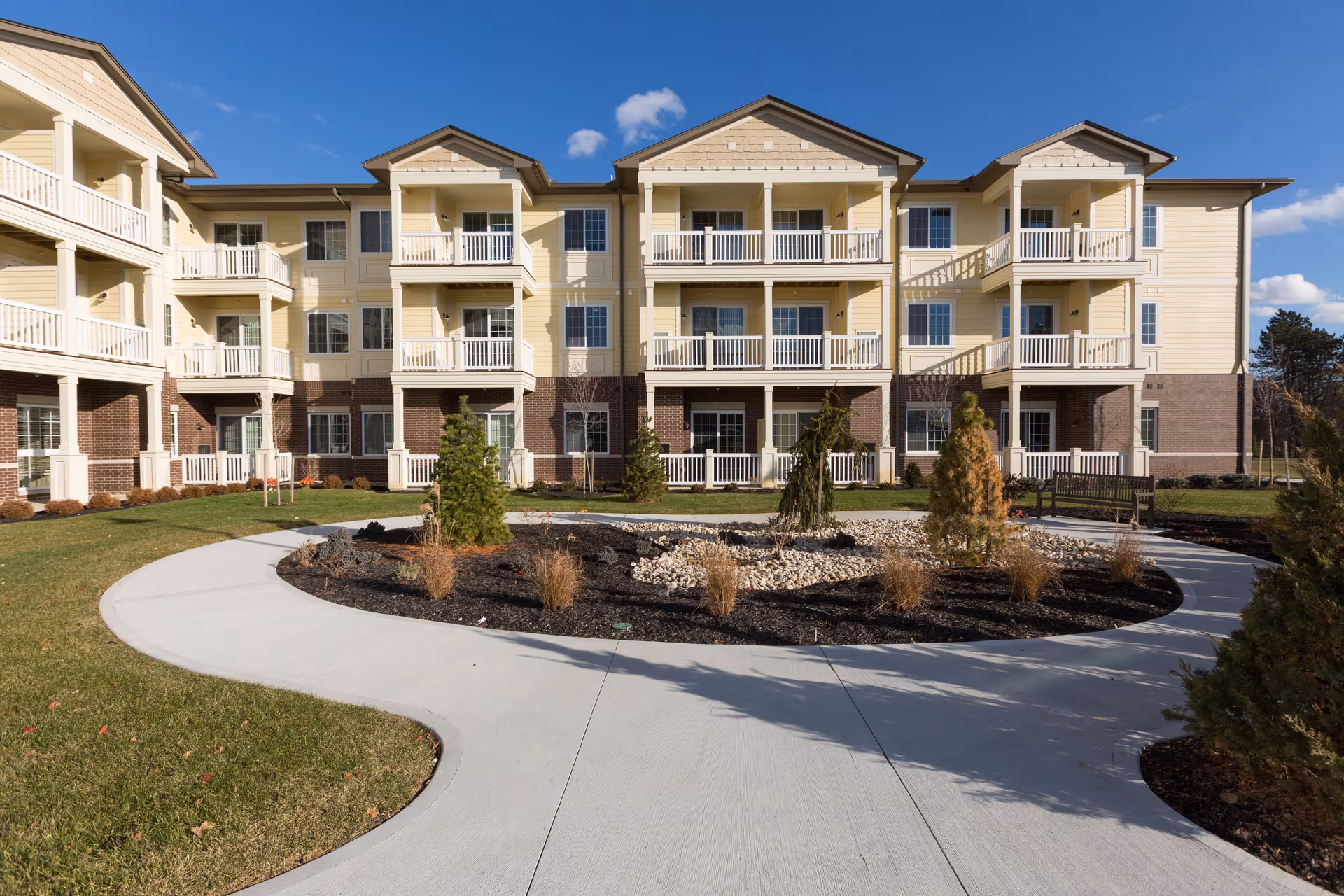 Exterior view of a three-story senior living facility building with balconies, surrounded by a landscaped garden area with a curved concrete walkway and small trees and shrubs under a clear blue sky.