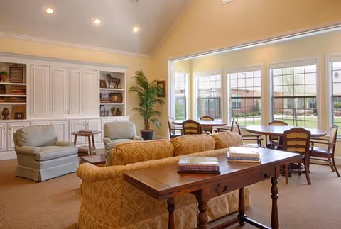 A bright and spacious living room area with large windows overlooking a garden. The room features a patterned sofa, two upholstered armchairs, a wooden table with books on it, and a dining table with chairs near the windows. Built-in white cabinetry and shelves with decorative items are visible along one wall, and a tall potted plant adds greenery to the space.
