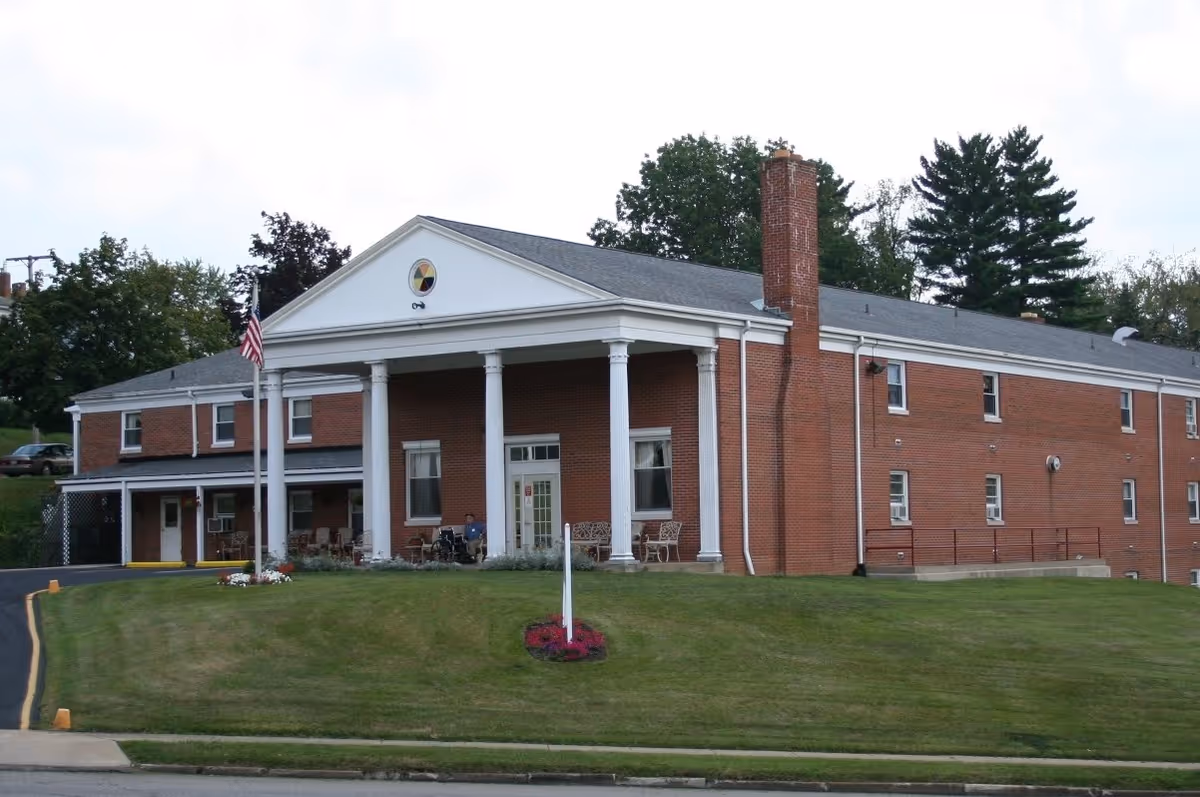 Exterior view of a two-story brick building with white columns at the entrance, a chimney, and an American flag on a flagpole in front. The building is surrounded by a well-maintained lawn with some flowers near the entrance and trees in the background.