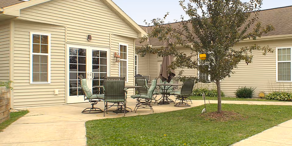 Outdoor patio area at a senior living facility with several metal tables and chairs with green cushions, a closed umbrella, a small tree, and beige siding building with white-framed windows and double glass doors.