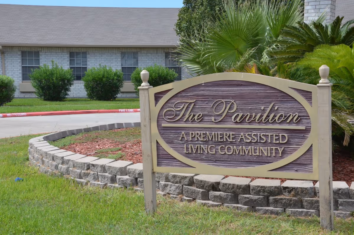 Outdoor view of a sign for The Pavilion, a premiere assisted living community, with a building and greenery in the background.
