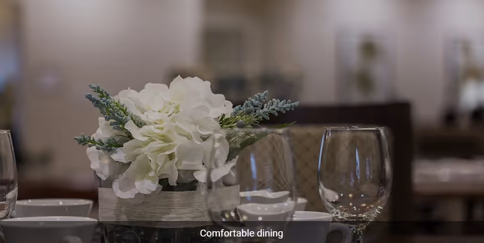 Close-up of a dining table set with a centerpiece of white flowers and greenery in a glass vase, surrounded by empty wine glasses and white cups in a softly lit dining room.