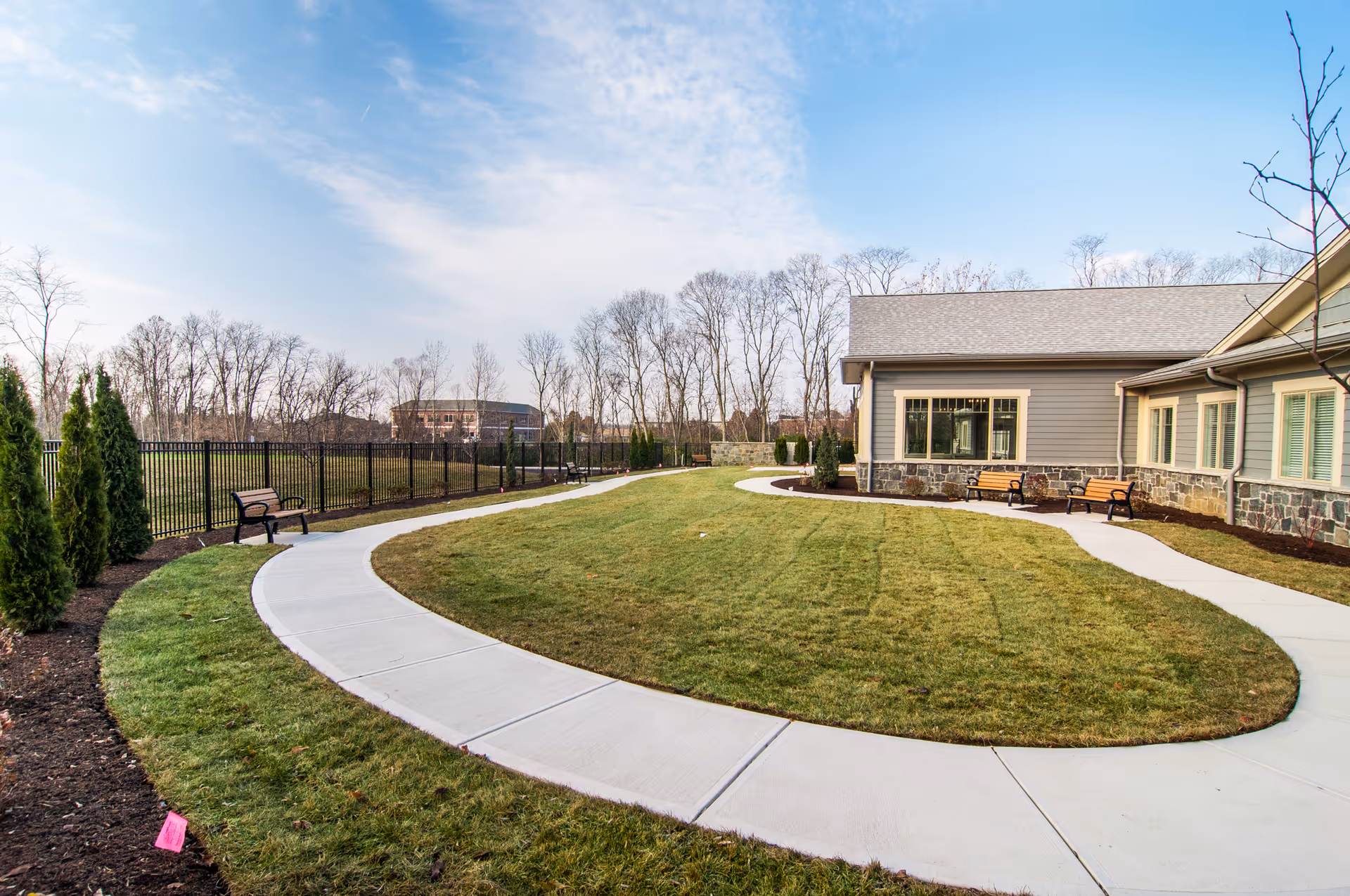 Curved concrete walkway surrounding a grassy area with benches along the path next to a single-story building with stone and siding exterior, under a partly cloudy sky.