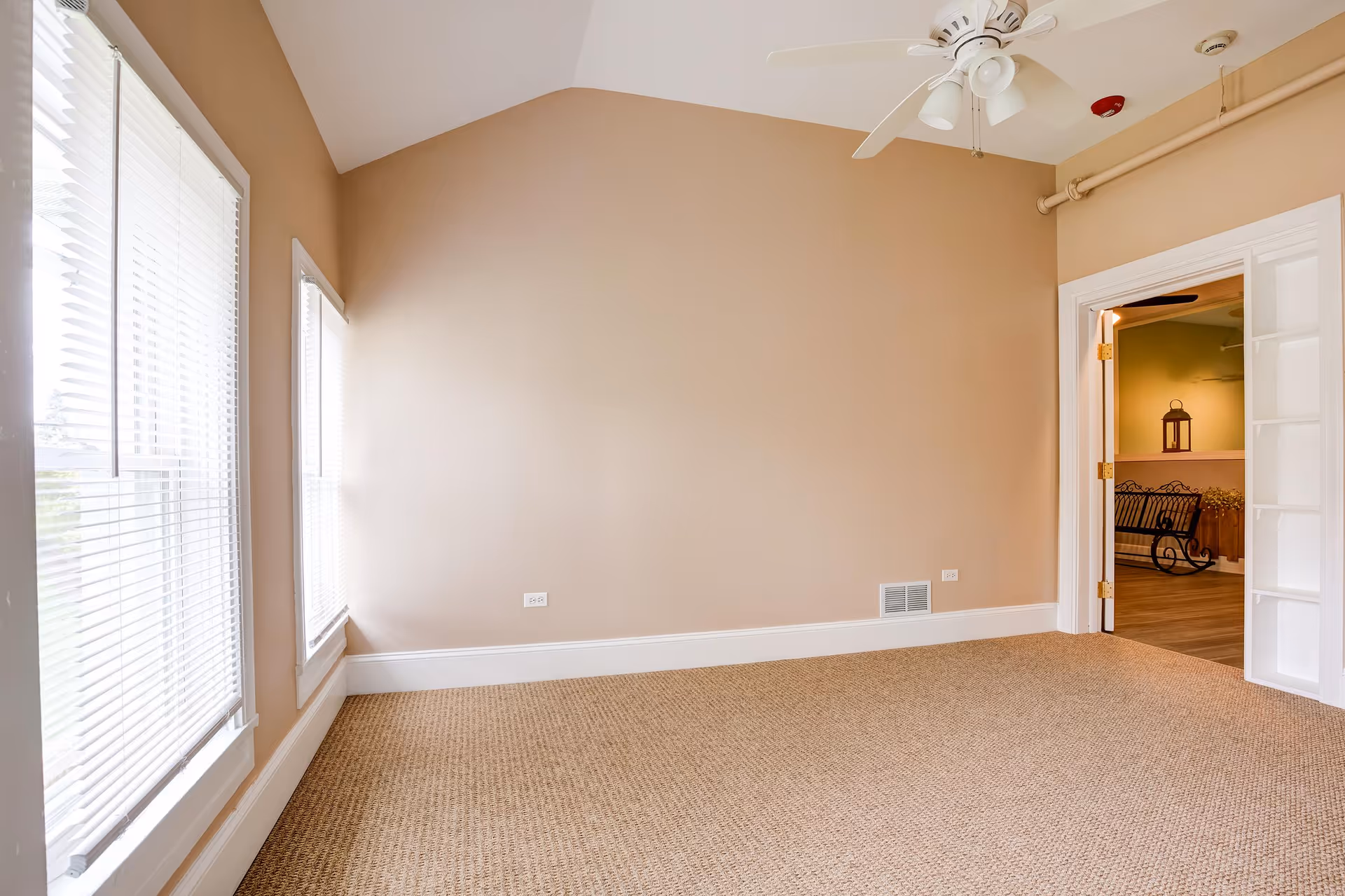 Empty room with beige walls, carpeted floor, two large windows with white blinds on the left, a white ceiling fan, and an open doorway leading to another room with a bench and decorative lantern.