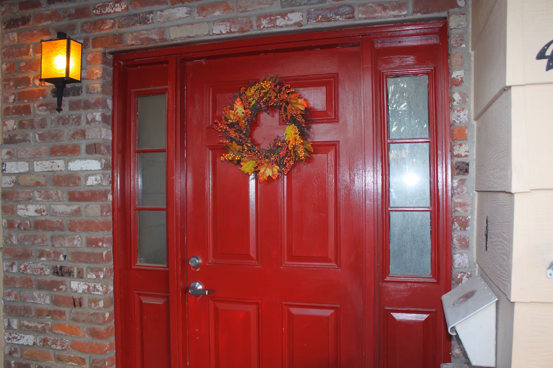 A bright red front door with decorative glass panels on each side and a fall-themed wreath hanging in the center. The door is set in a brick wall with a lit wall-mounted lantern on the left side and a mailbox on the right.