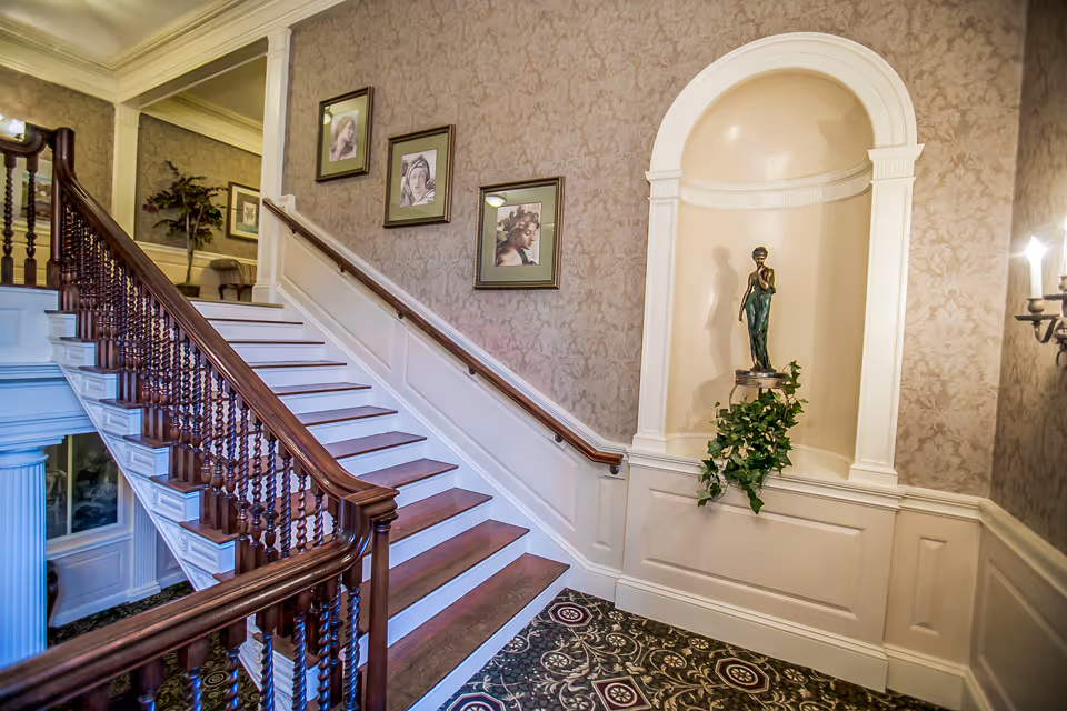 Interior view of a staircase with wooden handrails and white steps. The wall beside the staircase is decorated with framed artwork and patterned wallpaper. There is a niche in the wall with a statue of a woman and some greenery below it. The floor has a patterned carpet, and a wall sconce with candles is visible on the right side.