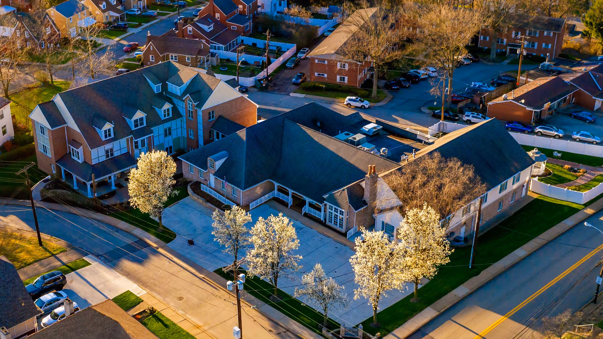 Aerial view of a residential neighborhood featuring a large building complex with multiple roofs and surrounding trees with white blossoms. The complex is adjacent to a street with parked cars and sidewalks, and other residential buildings are visible in the background.