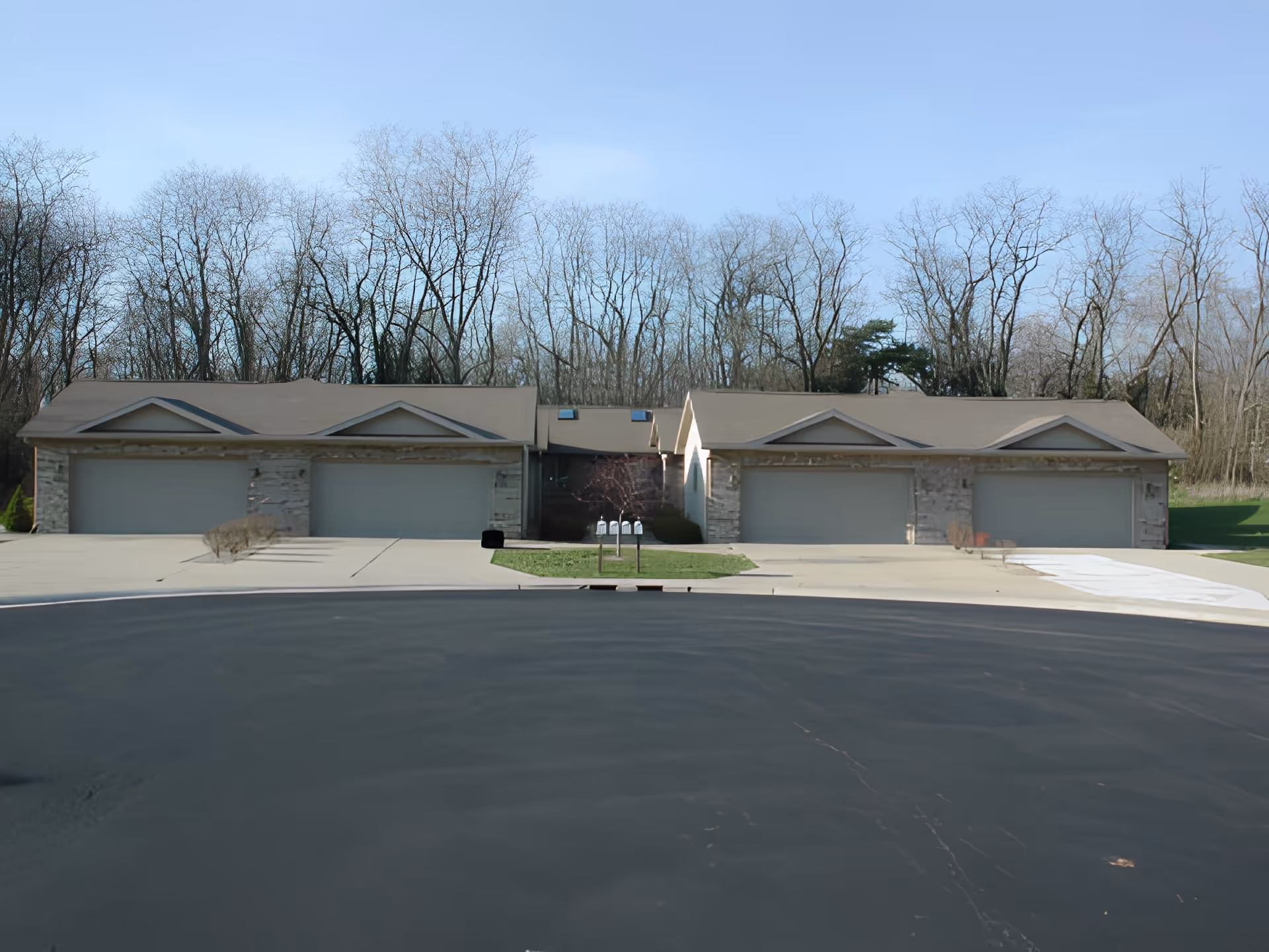 Exterior view of a senior living facility showing two connected buildings with multiple garage doors, a paved driveway, and leafless trees in the background under a clear blue sky.