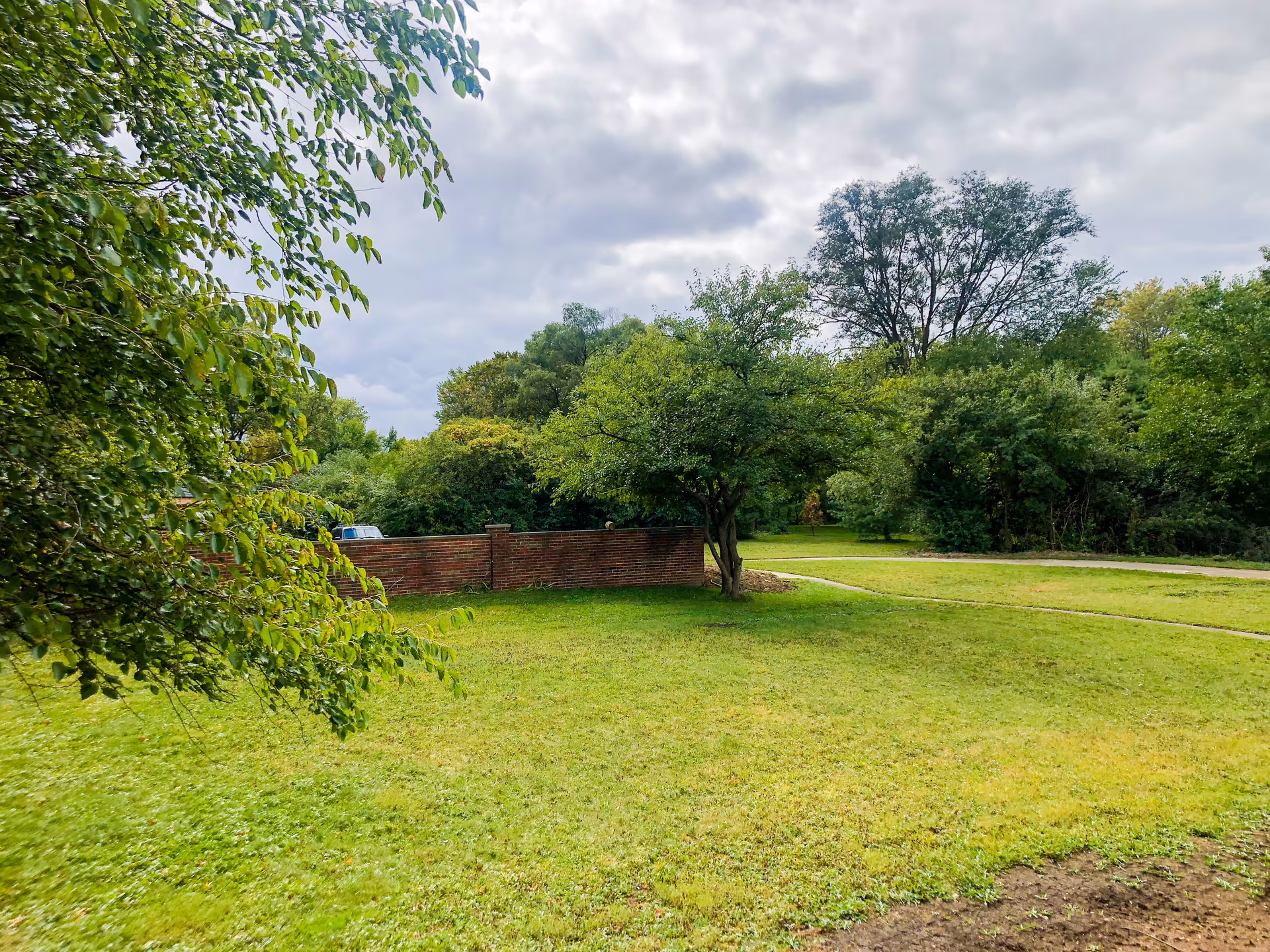 A grassy outdoor area with a few trees and a brick wall in the background under a cloudy sky.