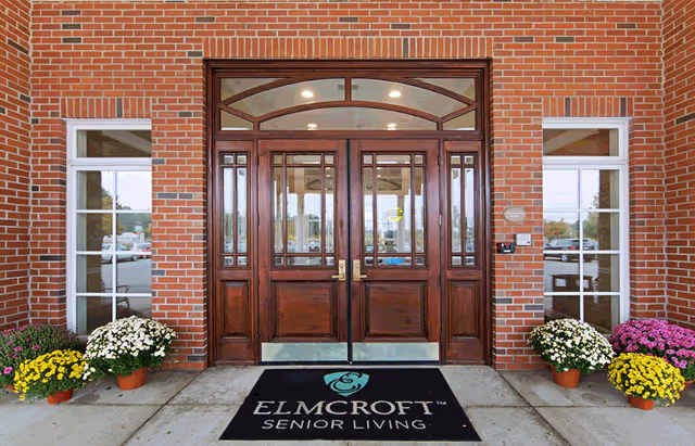 Entrance of a senior living facility with double wooden doors framed by brick walls and windows on either side. There are potted flowers in yellow, white, and purple colors placed on both sides of the entrance. A black mat in front of the doors displays the ElmCroft Senior Living logo.