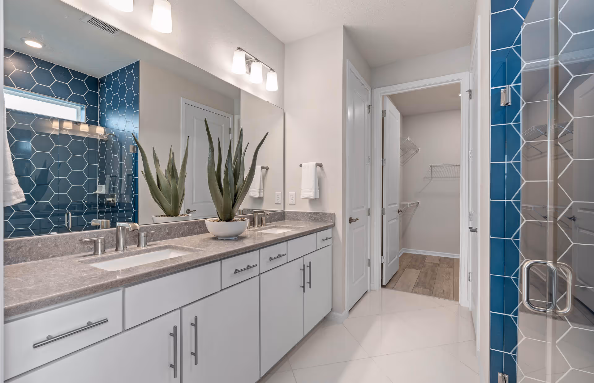 Modern bathroom with a double sink vanity featuring gray countertops and white cabinets. A large mirror spans the wall above the sinks with two light fixtures mounted above. To the right, there is a glass-enclosed shower with blue hexagonal tiles. In the background, an open door reveals a walk-in closet with wooden flooring and wire shelving.