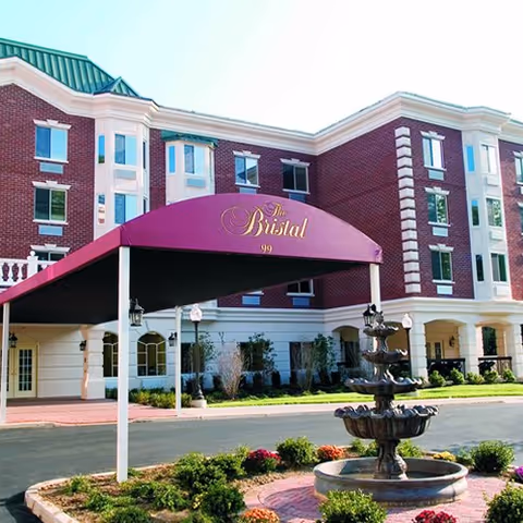 Exterior view of The Bristal Assisted Living at North Hills building with a red brick facade, white trim, and a green roof. A maroon canopy with the facility name and number 99 is at the entrance. In front of the building is a multi-tiered water fountain surrounded by landscaping and flowers.