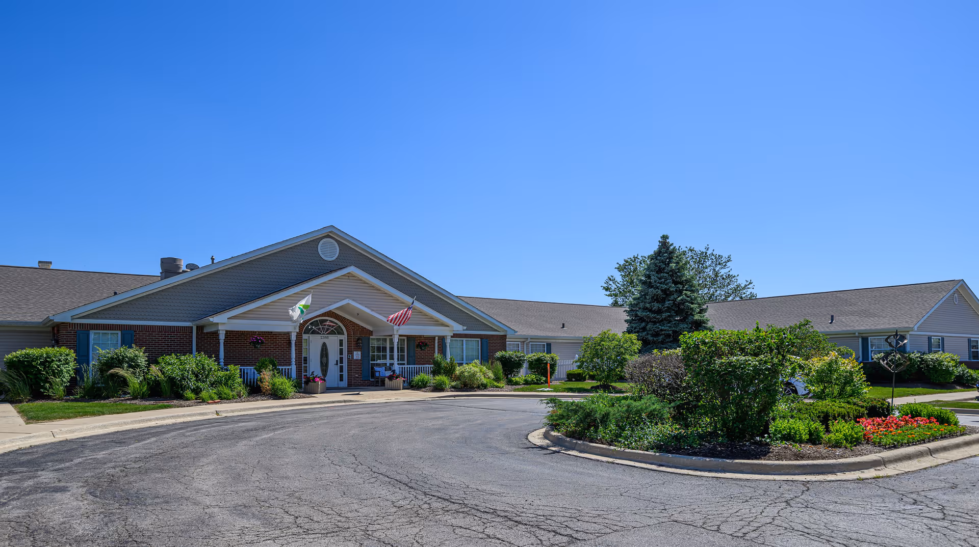 Exterior view of Arden Courts A ProMedica Memory Care Community in Geneva, showing a single-story building with a peaked roof, an entrance with a covered porch, two flags, and well-maintained landscaping including bushes, flowers, and a tree in a circular driveway.