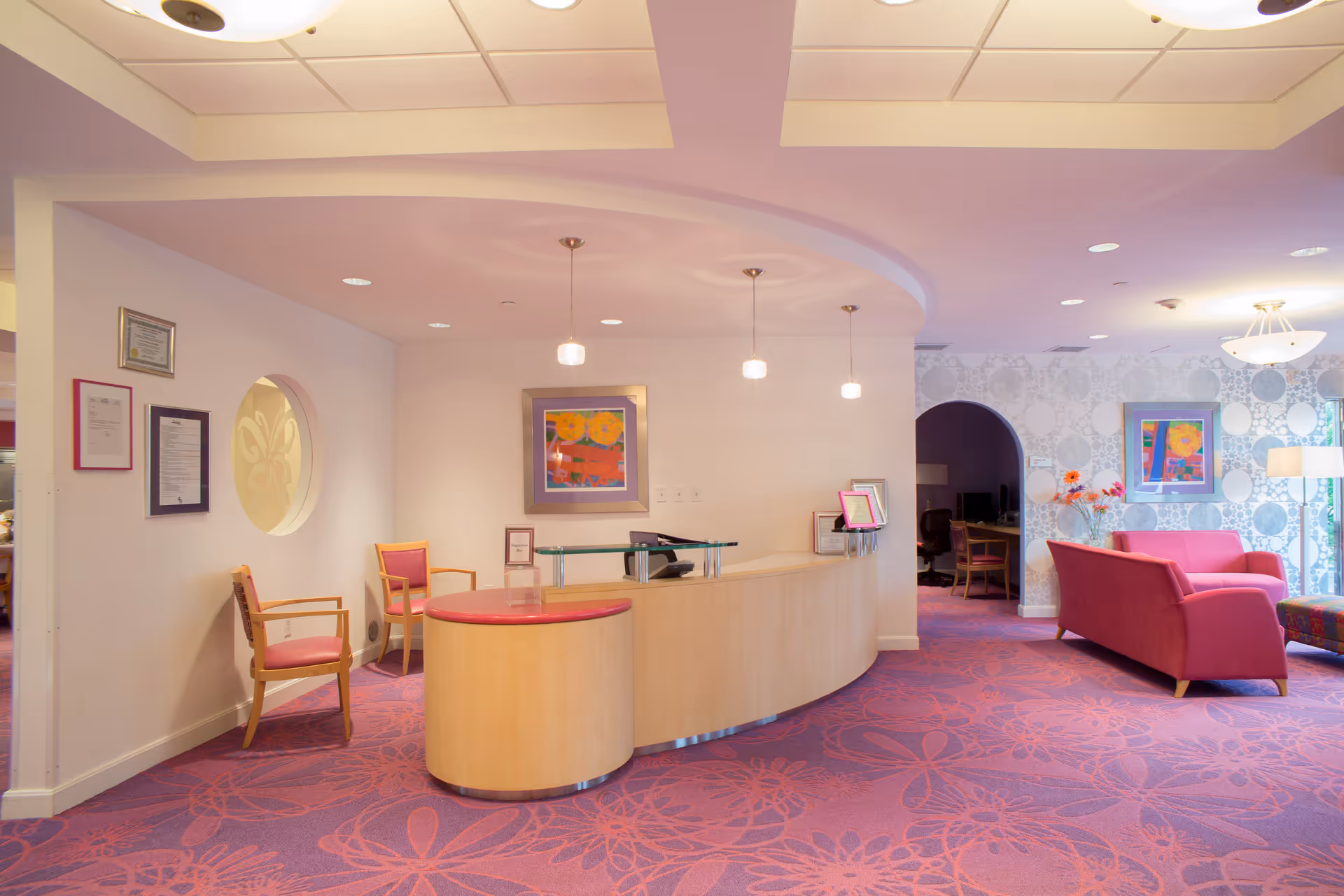 Reception area with a curved wooden desk and three hanging pendant lights above it. There are two wooden chairs with pink cushions to the left of the desk. The floor is covered with a purple carpet featuring a floral pattern. To the right, there is a seating area with pink couches, a floor lamp, and colorful artwork on the walls. The walls are light-colored with a circular patterned wallpaper in the seating area.