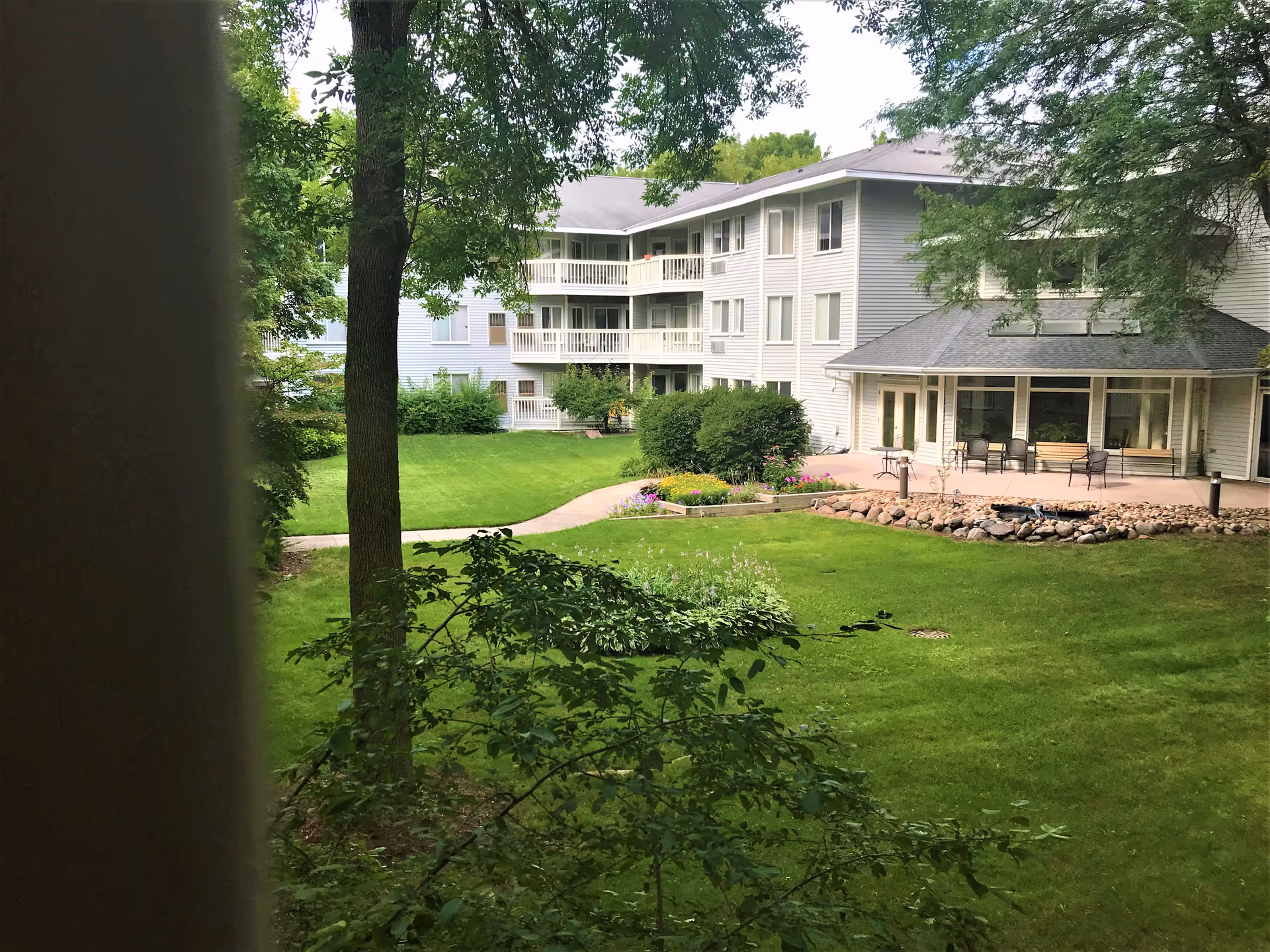 View of a senior living facility's outdoor courtyard with green grass, trees, bushes, and a paved walkway. The building has multiple floors with balconies and large windows. There is a patio area with chairs and a fire pit surrounded by rocks.