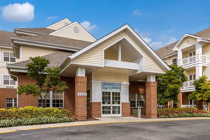 Front exterior view of a senior living facility building with a covered entrance supported by brick columns, surrounded by well-maintained landscaping including trees and flower beds under a blue sky with a few clouds.