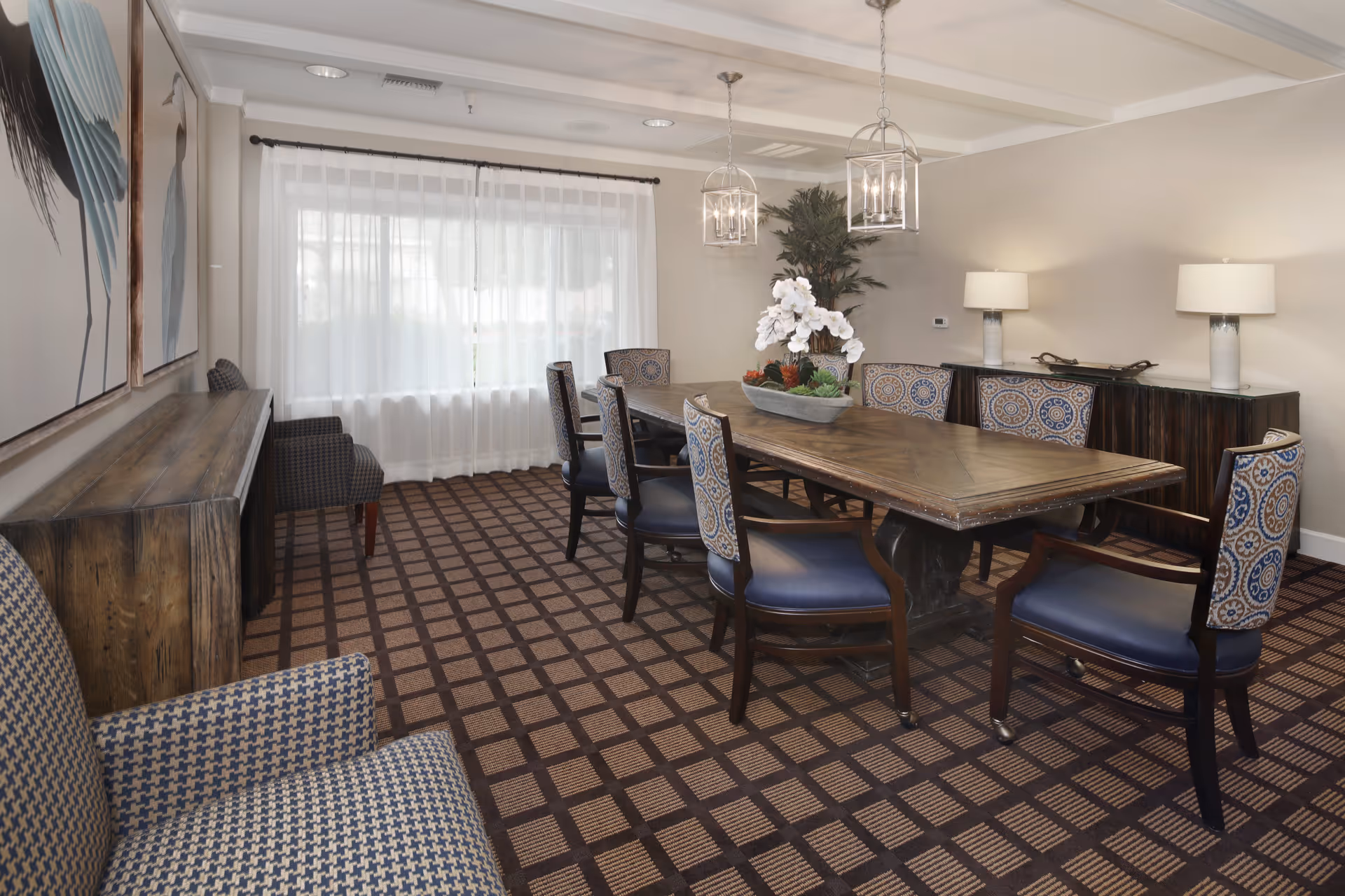 A well-lit dining room with a long wooden table surrounded by eight upholstered chairs with patterned backs and blue seats. The room features a patterned carpet, two hanging lantern-style light fixtures, a large window with sheer white curtains, two table lamps on a dark wooden sideboard, and decorative artwork on the walls.
