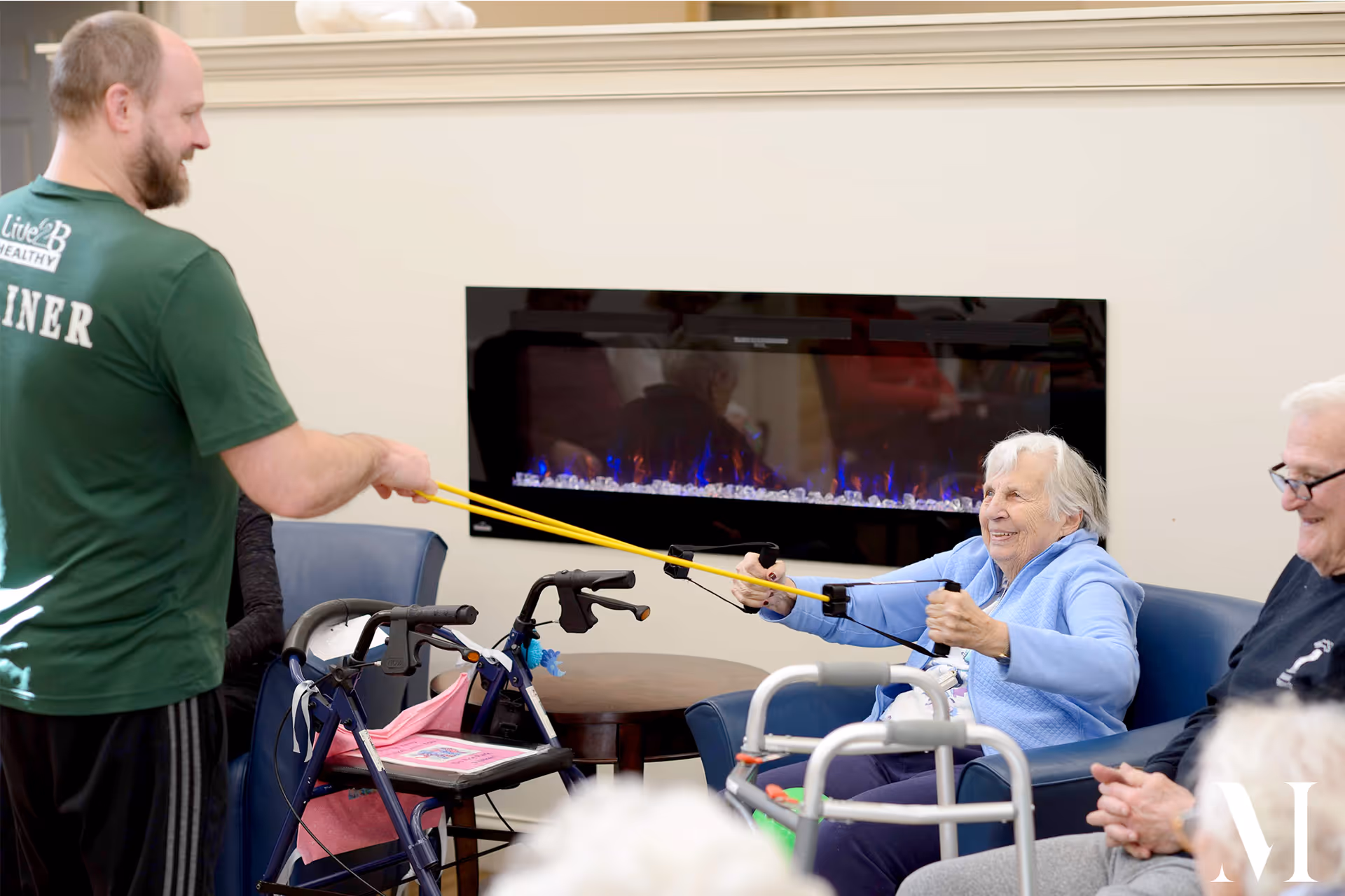 An elderly woman seated in a blue armchair is exercising with a resistance band held by a male trainer wearing a green shirt. Other elderly individuals are seated nearby, and there is a modern electric fireplace mounted on the wall behind them.