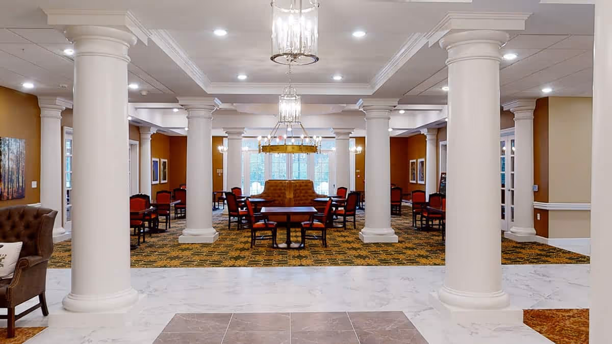 Spacious senior living facility common area with large white columns, patterned carpet, multiple tables and chairs with red upholstery, and large windows letting in natural light. The ceiling features recessed lighting and hanging chandeliers.