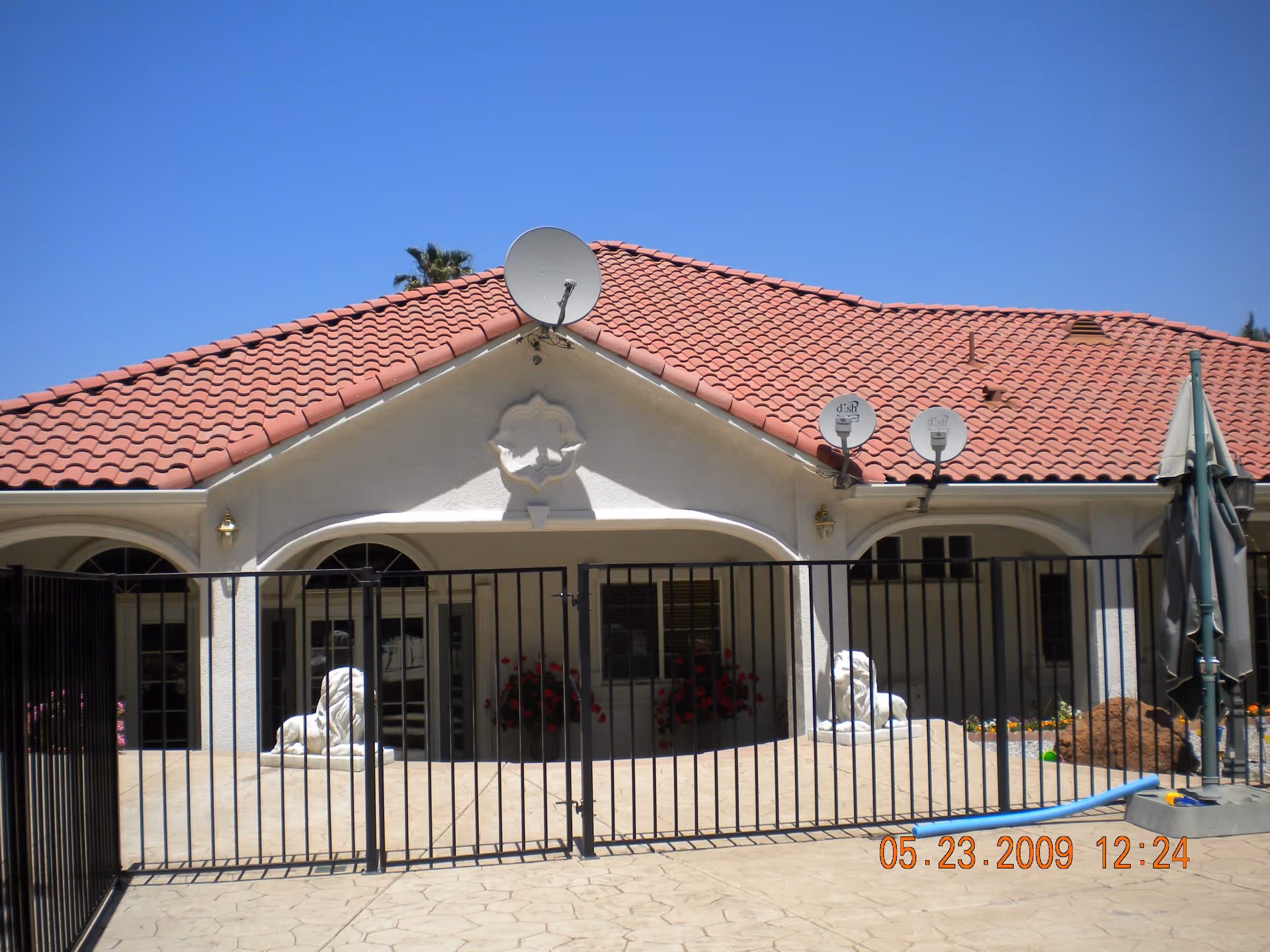 Exterior view of a single-story building with a red tiled roof and white walls, featuring three satellite dishes mounted on the roof. The building has arched doorways and windows, with two white lion statues placed behind a black metal fence. There are some potted plants with red flowers visible near the entrance, and a closed green umbrella on the right side. The sky is clear and blue.