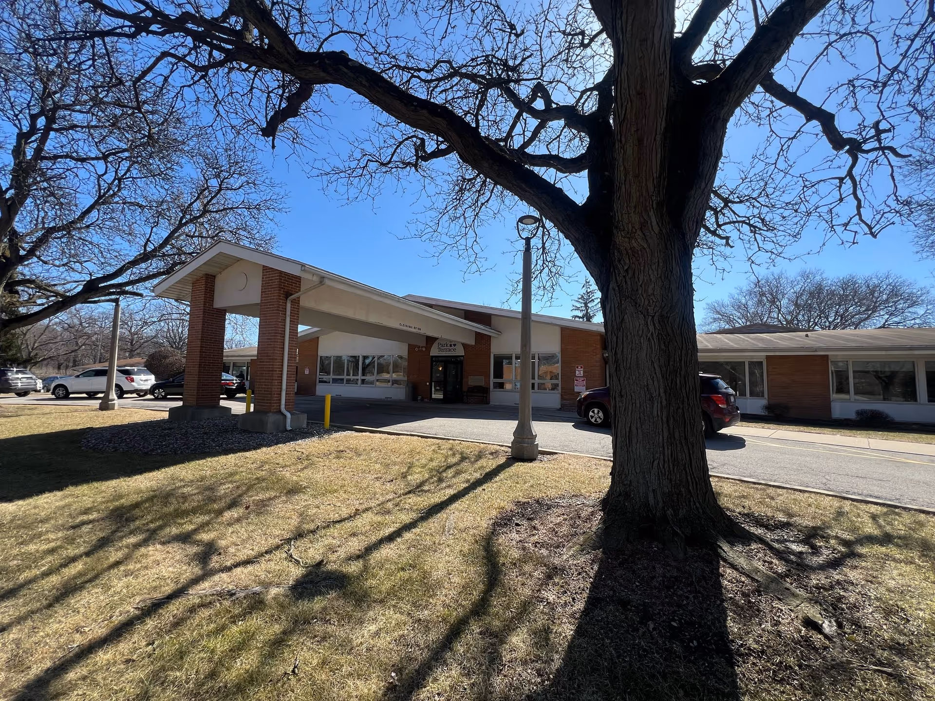 Exterior view of Park Terrace Rehabilitation Center on a sunny day with a clear blue sky. The building has a brick facade with a covered entrance supported by brick columns. Several cars are parked near the entrance, and leafless trees cast shadows on the grassy area in front.