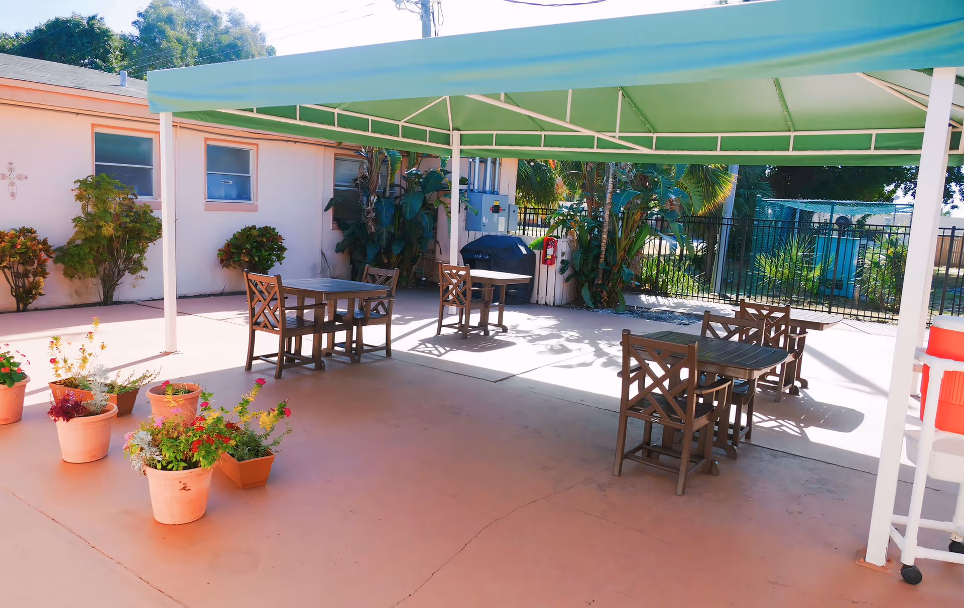 Outdoor patio area with a green canopy providing shade over several wooden tables and chairs. There are potted plants with flowers on the concrete floor, and a building wall with windows and some greenery in the background. A black grill and a red cooler are also visible near the back fence.