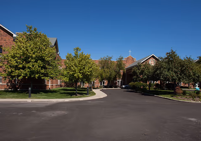 Exterior view of a brick building with a cross on the roof, surrounded by green trees and a paved driveway under a clear blue sky.