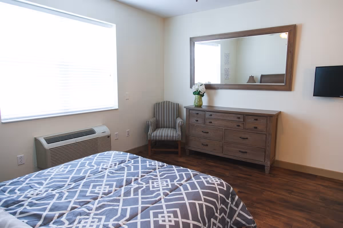 A bedroom with a bed covered in a gray and white geometric patterned comforter, a wooden dresser with multiple drawers, a large rectangular mirror above the dresser, a striped armchair in the corner, a wall-mounted TV, and a window with blinds allowing natural light to enter. The floor is wooden and the walls are painted white.